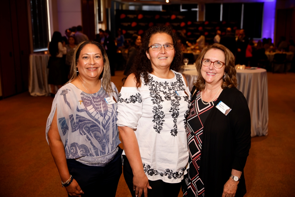 Three women pose together while reception guests gather in the background.